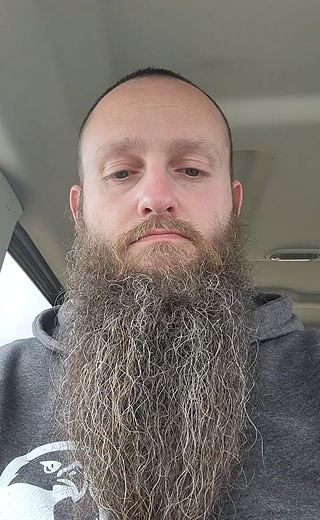A close-up selfie of a man with a very long, thick, wavy gray and brown beard, wearing a dark gray hoodie inside a vehicle.