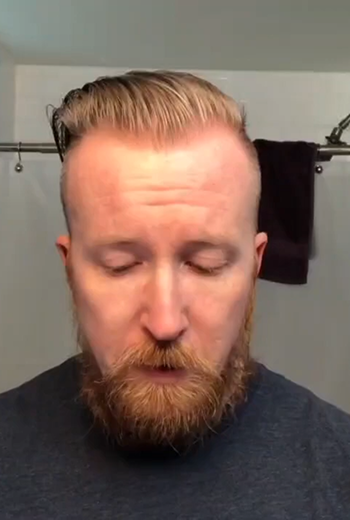 A close-up shot of a man with a blonde receding hairline and a reddish-brown beard, wearing a dark gray t-shirt. He is looking down, and a bathroom setting with a shower curtain and a dark towel is visible in the background.