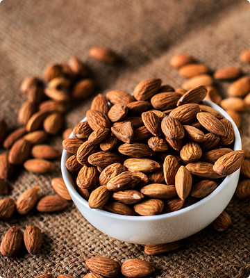 A white ceramic bowl filled with whole almonds, set on a rustic burlap surface with additional almonds scattered around.