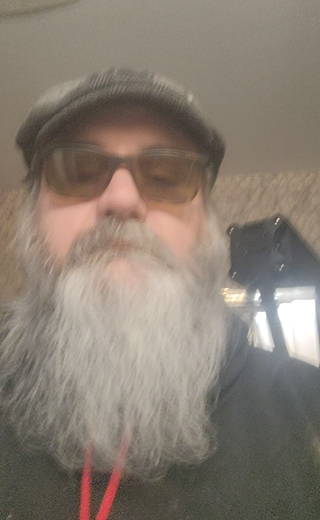 A close-up selfie of a man with a long gray beard, wearing a gray flat cap, tinted sunglasses, and a black hoodie with a red lanyard.